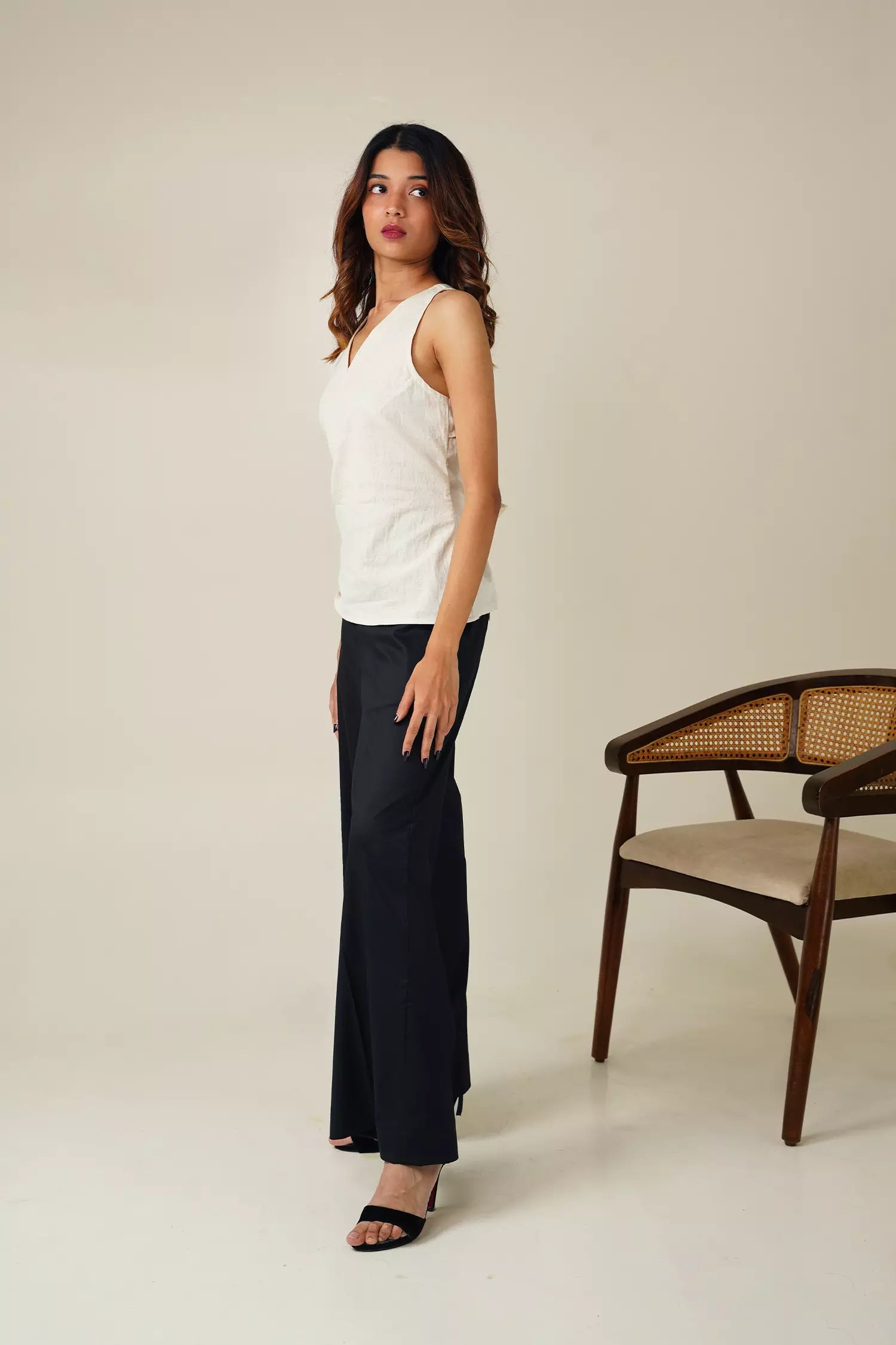 side view of Woman wearing a white sleeveless top with v neckline and black pants against a neutral background