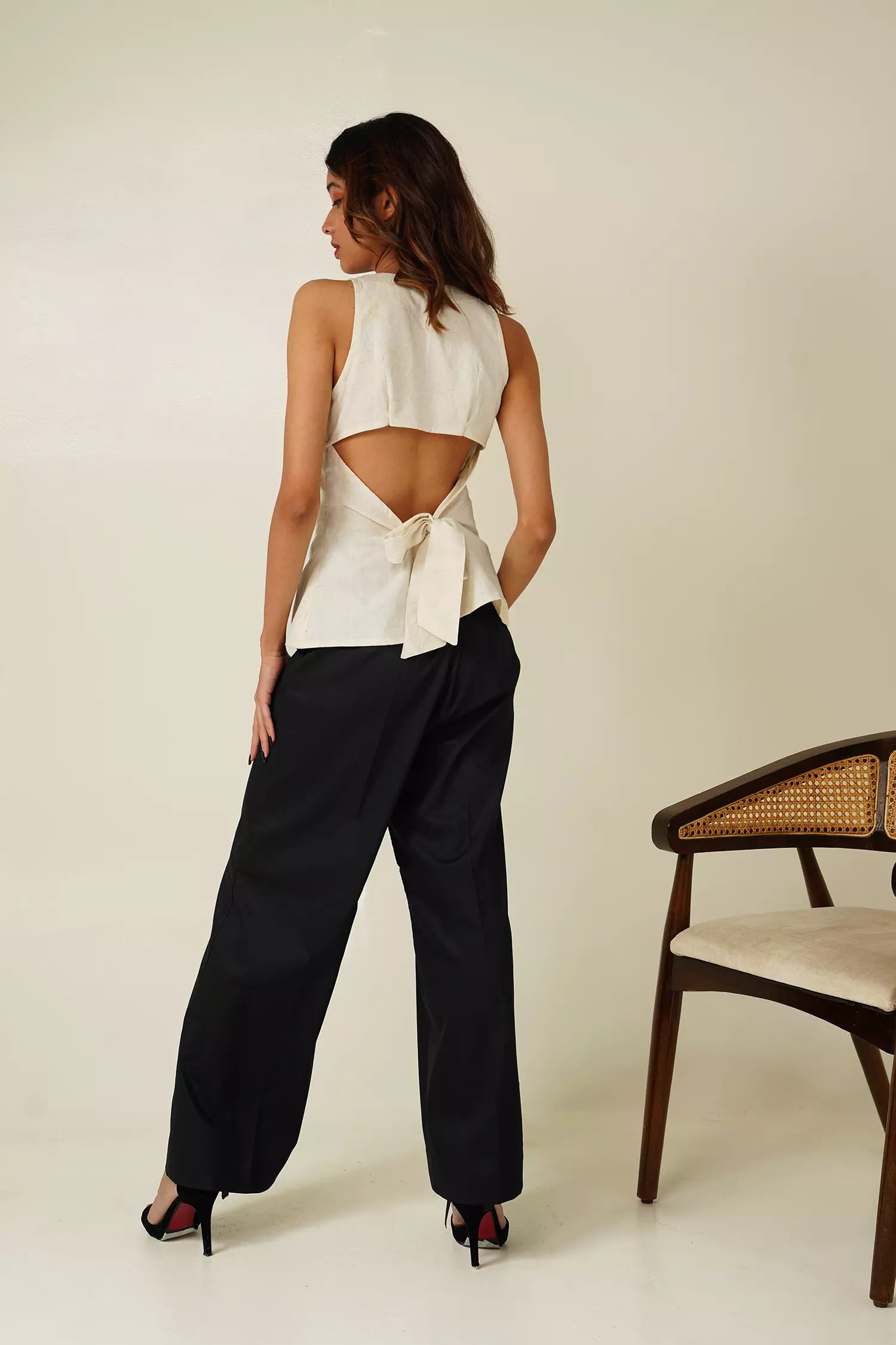 back view of Woman wearing a white shackles top with tie detail and black pants against a neutral background