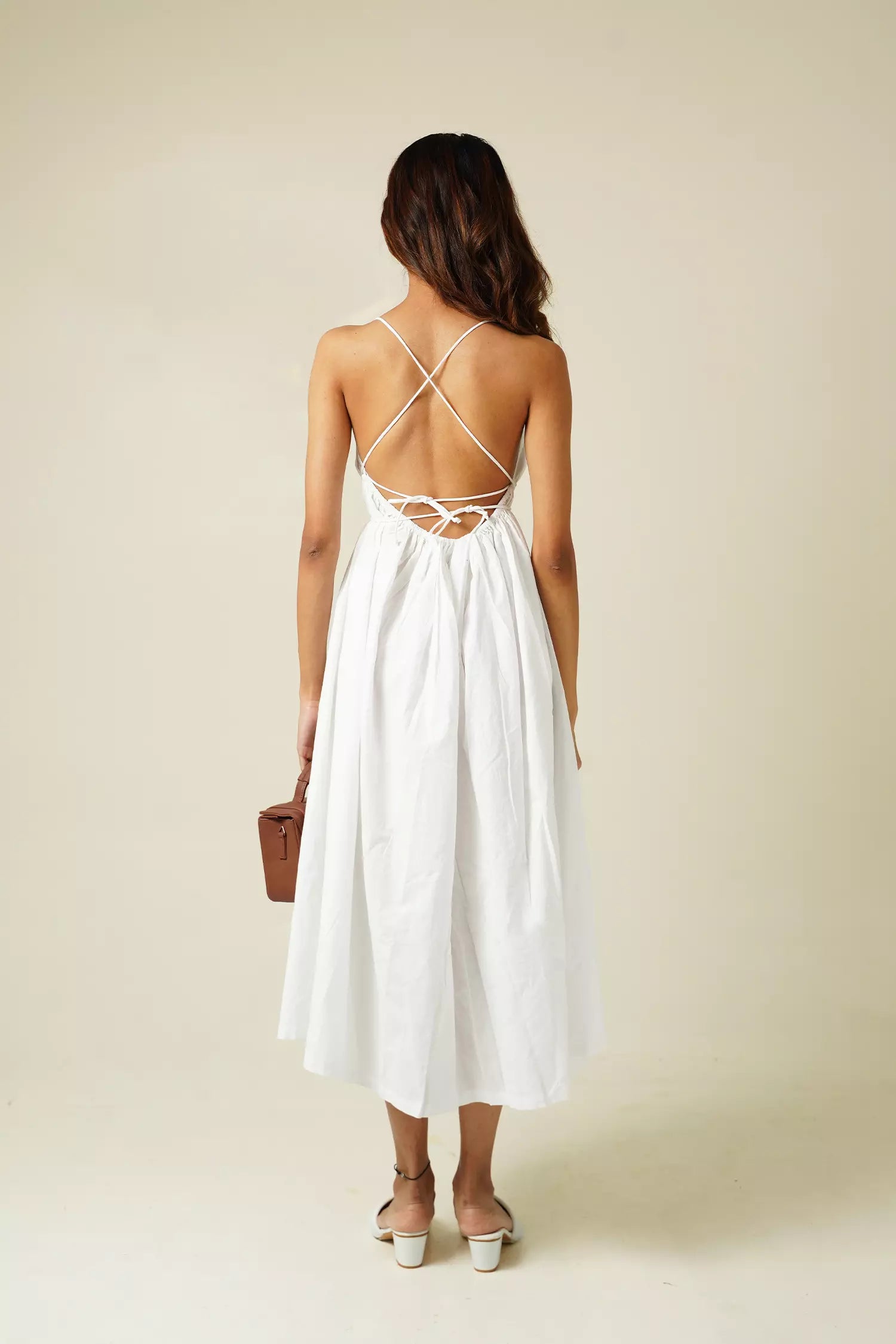 back view of Woman wearing a white cotton sleeveless midi dress with backless criss cross design against a plain background