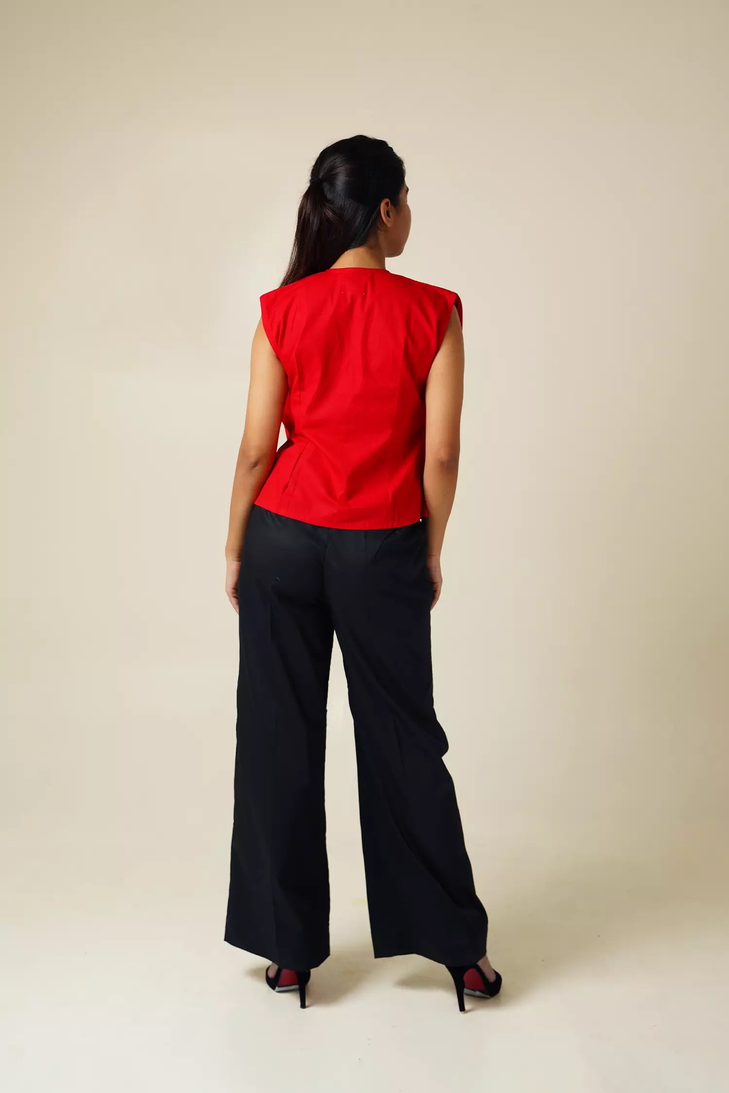 back view of Woman wearing a red pure cotton pleated top with plunging neckline and black pants against a beige background