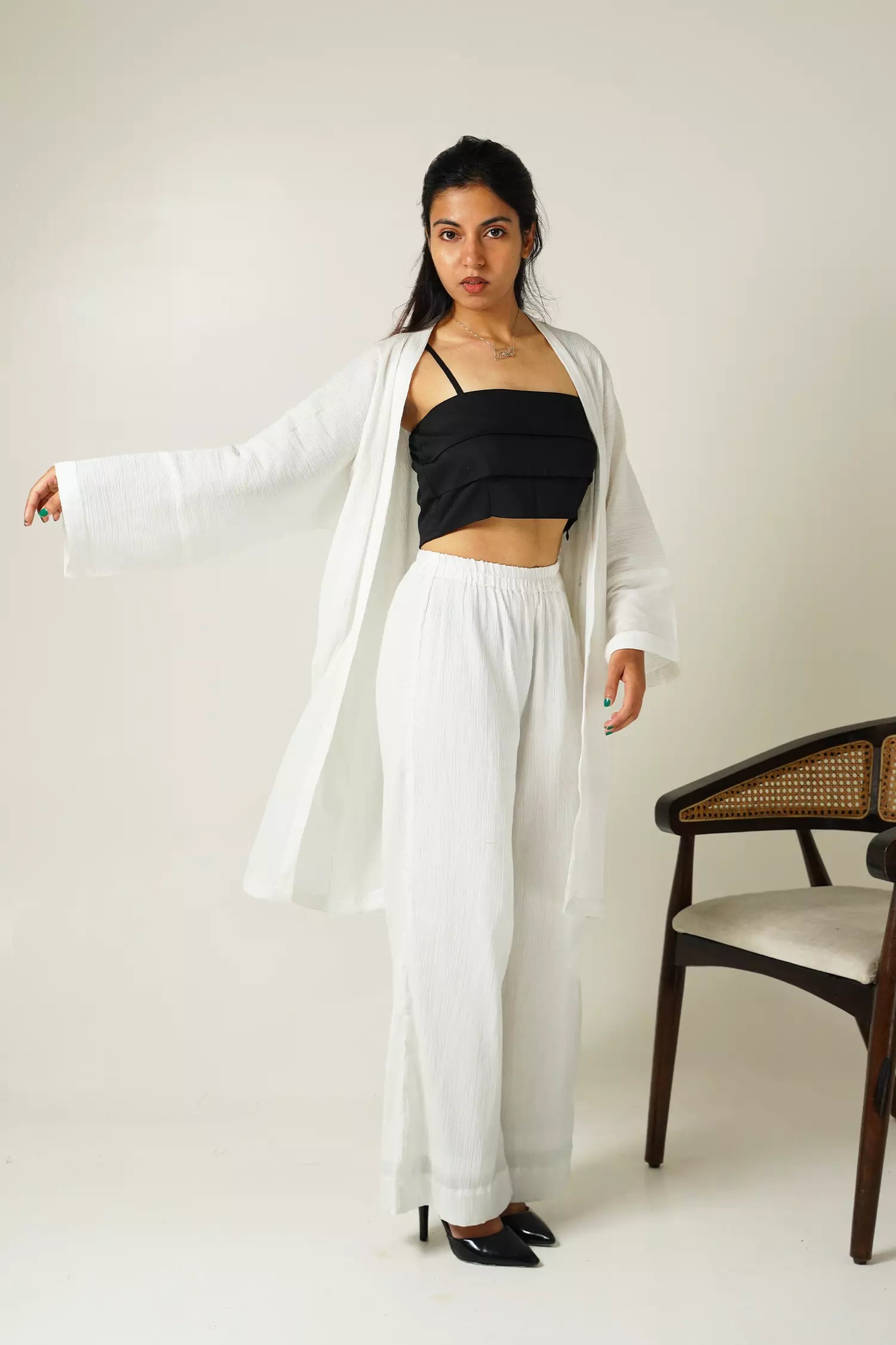 front view of Woman wearing a white two piece set of kaftan style overall jacket and wide fit pants with a black top, standing against a plain background.