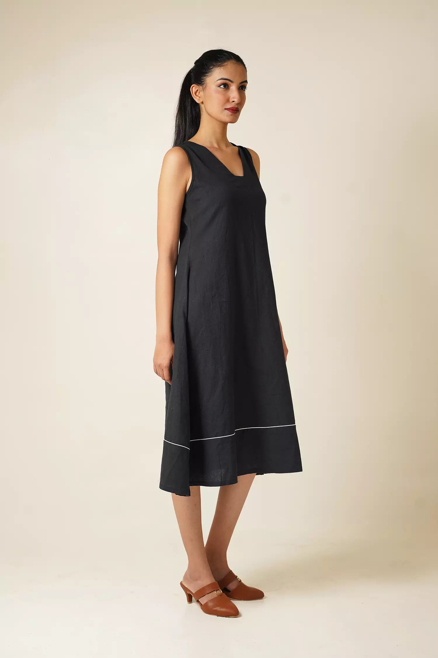 side view of Woman wearing a black knee length sleeveless dress with modified v neckline on a beige background