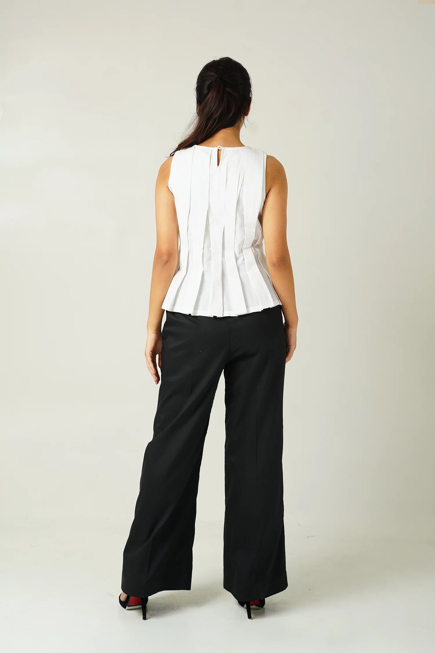 back view of Woman wearing a white sleeveless top with pleat details and black pants against a plain background