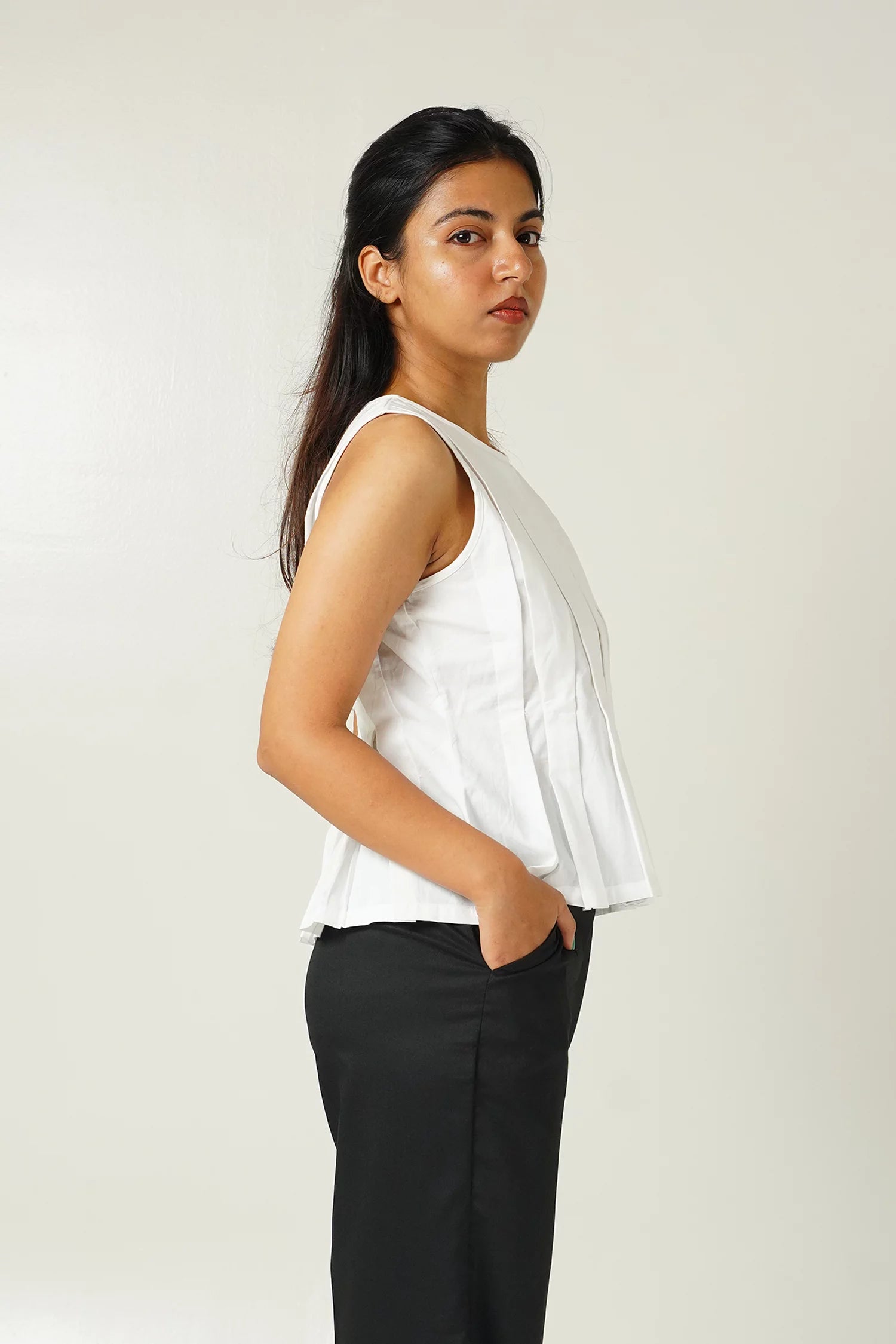 close up side view of Woman wearing a white sleeveless top with pleat details and black pants against a plain background