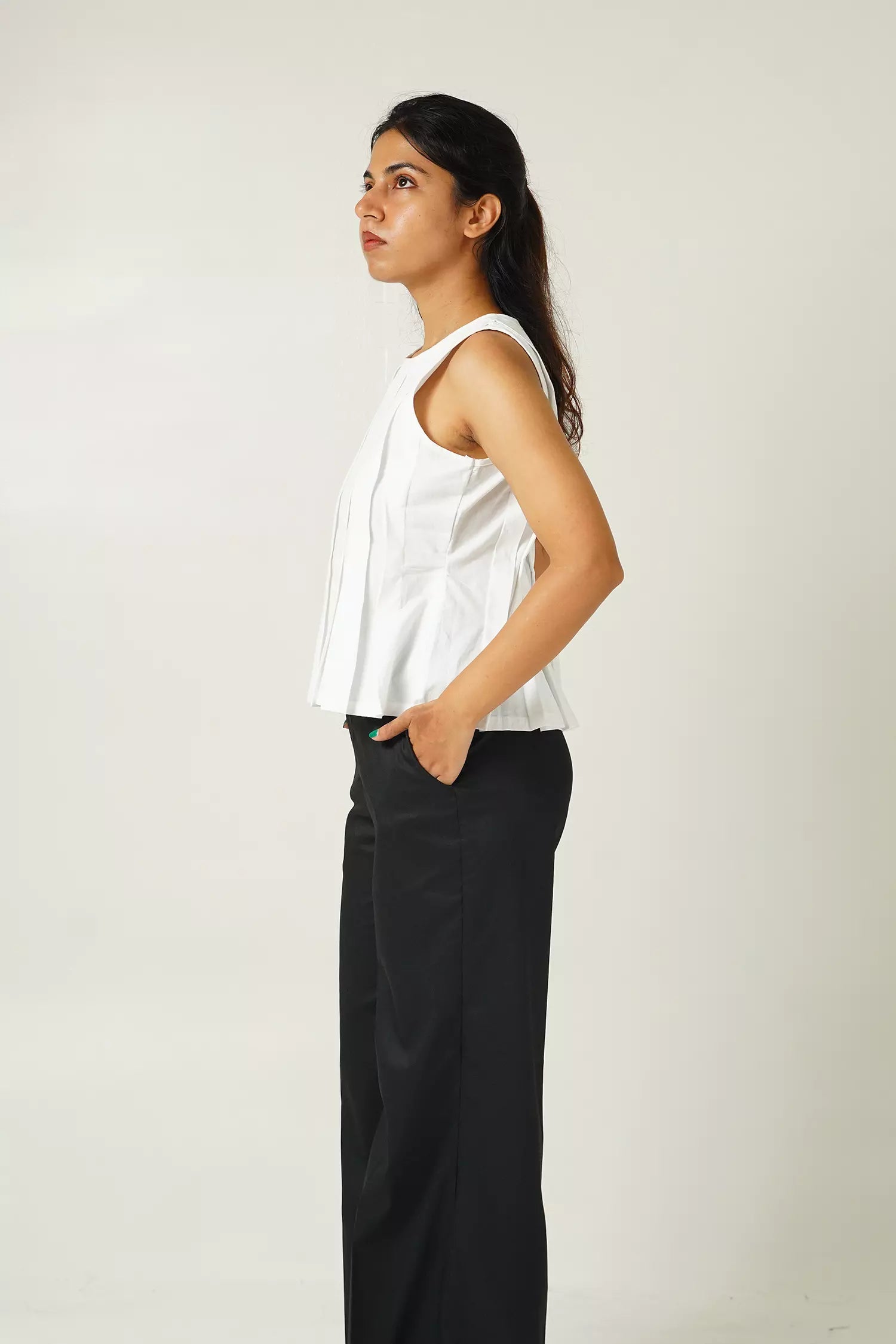 close up side view of Woman wearing a white sleeveless top with pleat details and black pants against a plain background