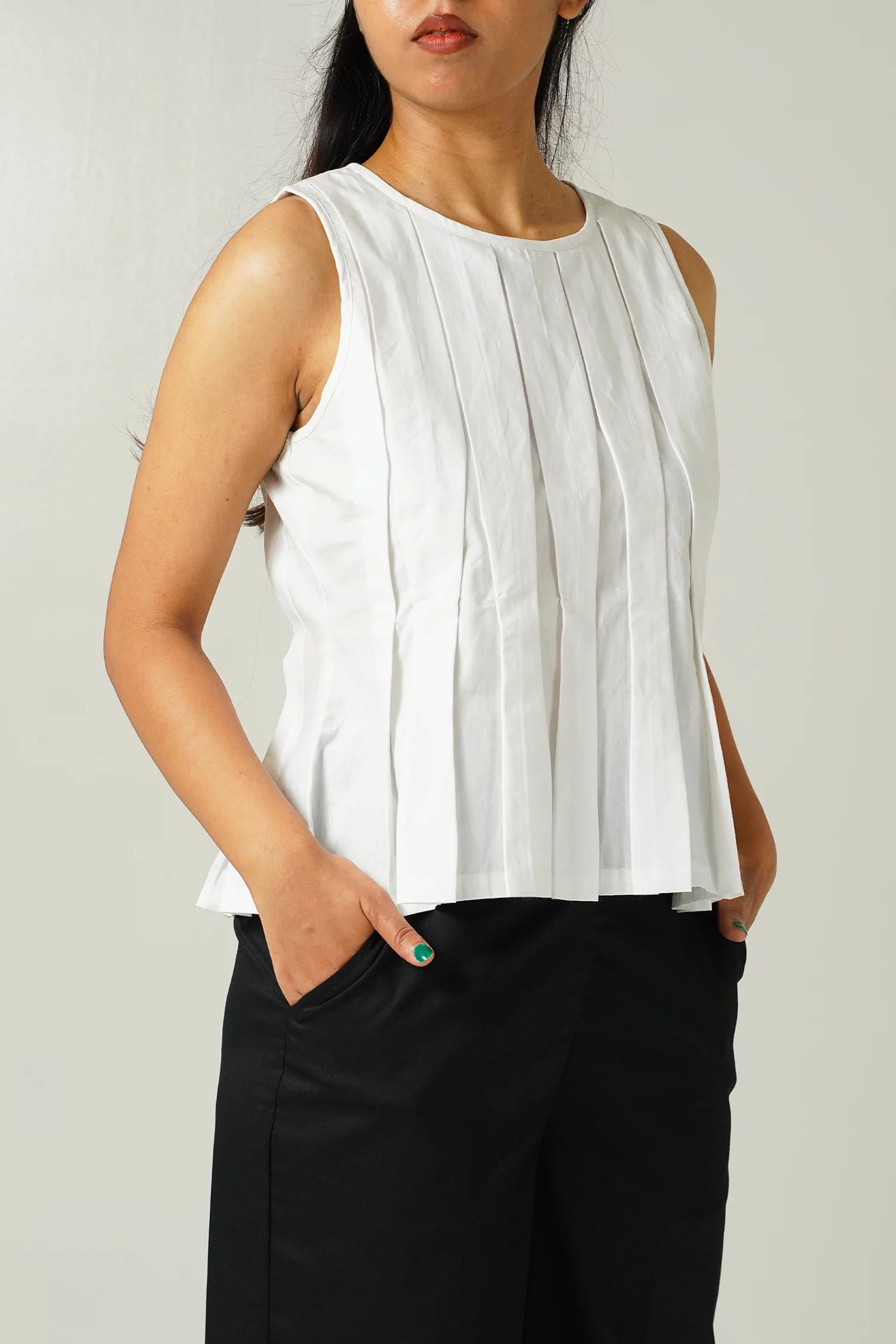 close up front view of Woman wearing a white sleeveless top with pleat details and black pants against a plain background
