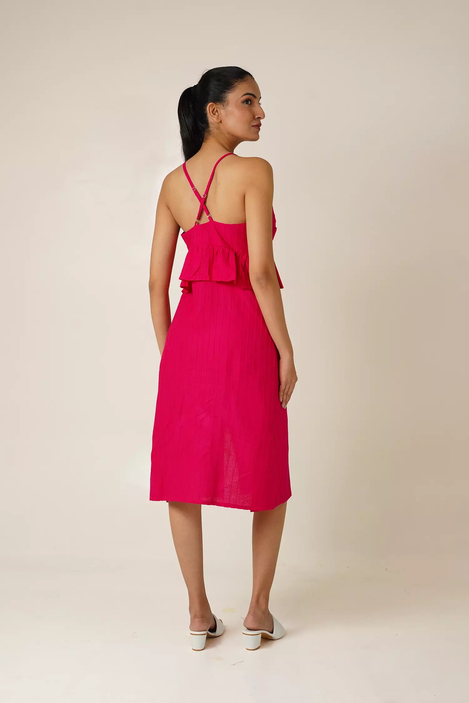 back view of Woman wearing a bright pink sleeveless and backless dress with a v neckline, peplum and a side slit on a beige background