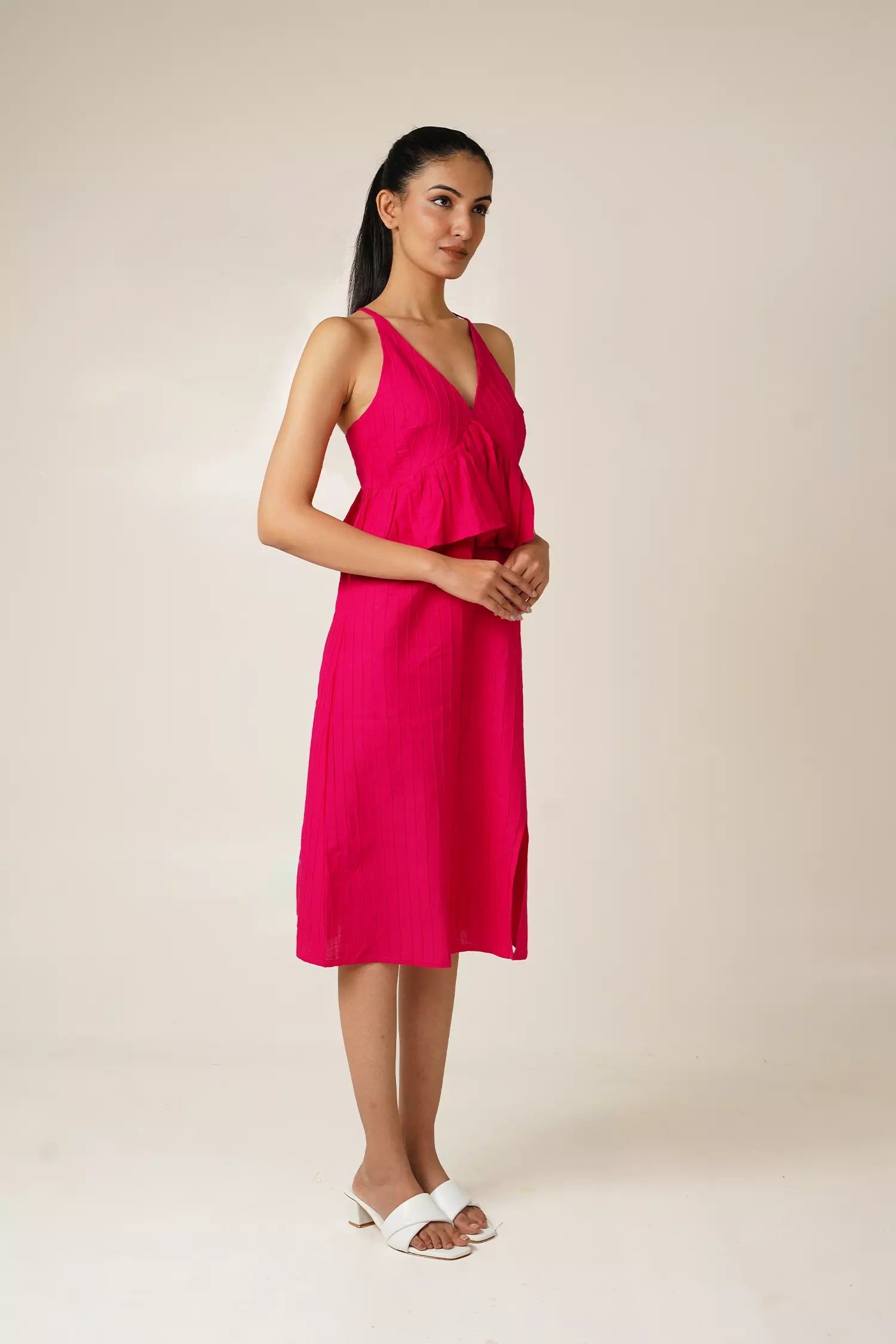 side view of Woman wearing a bright pink sleeveless and backless dress with a v neckline, peplum and a side slit on a beige background