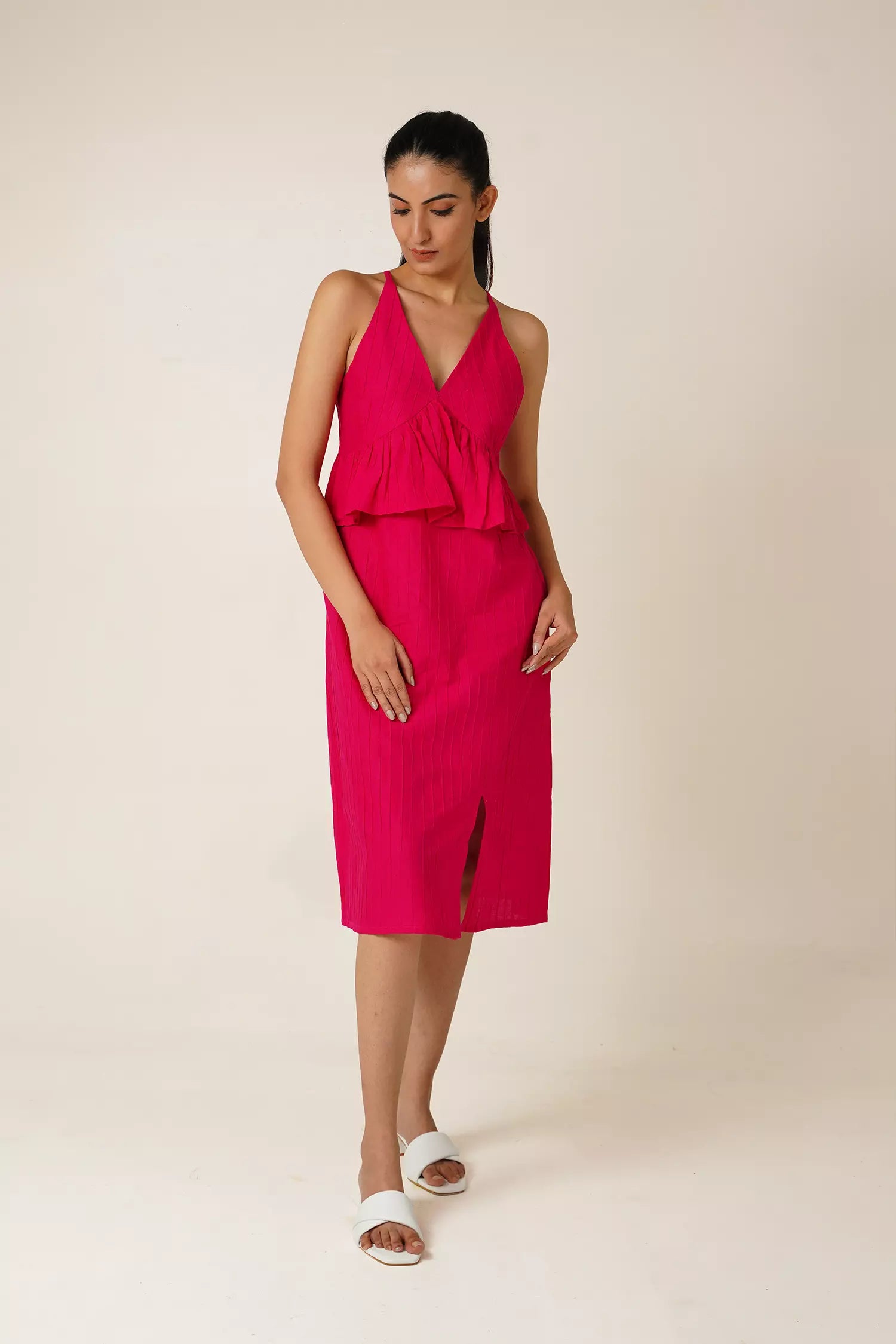 front view of Woman wearing a bright pink sleeveless and backless dress with a v neckline, peplum and a side slit on a beige background