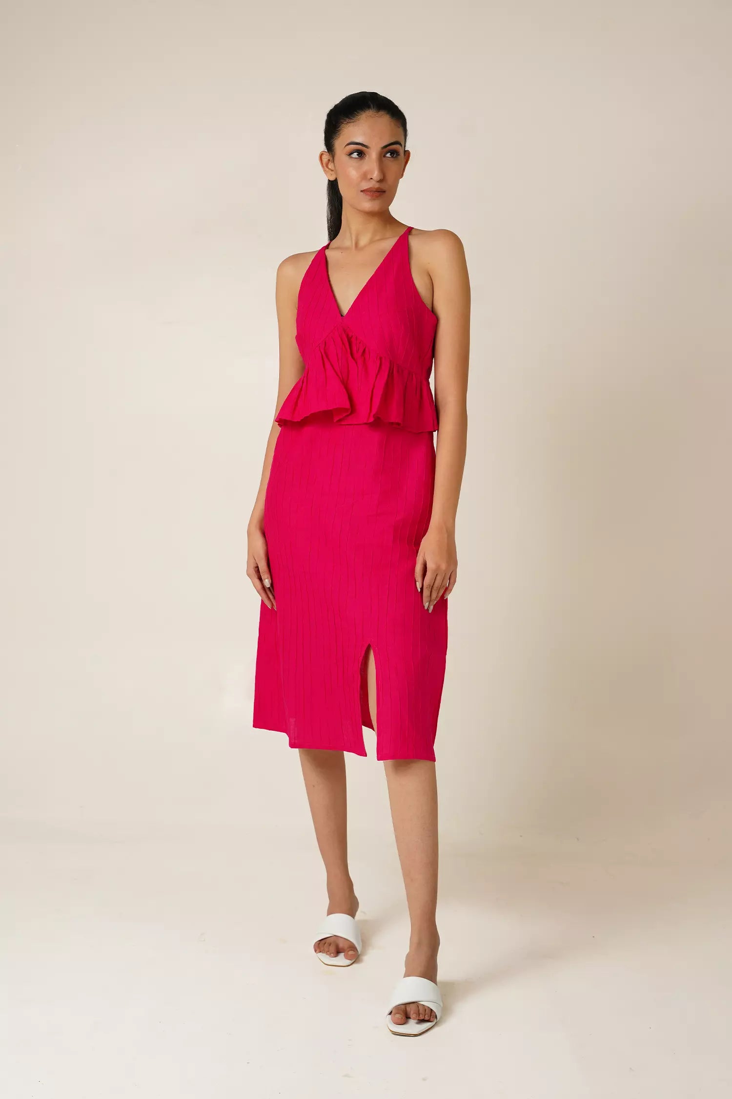 front view of Woman wearing a bright pink sleeveless and backless dress with a v neckline, peplum and a side slit on a beige background