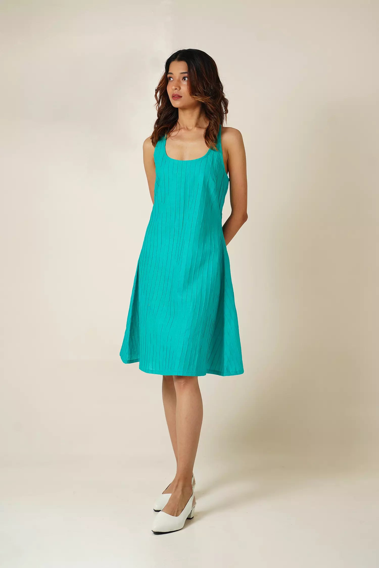 front view of Woman wearing a teal sleeveless knee length dress with pin tucks on cotton fabric against a beige background