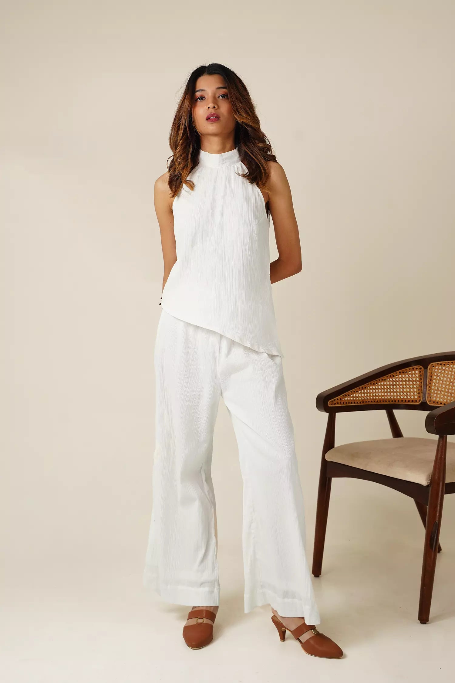 Woman wearing a white sleeveless halter neck top with asymmetrical hemline and wide-leg pants in white crinkle white cotton fabric standing in a minimalistic setting with a chair.