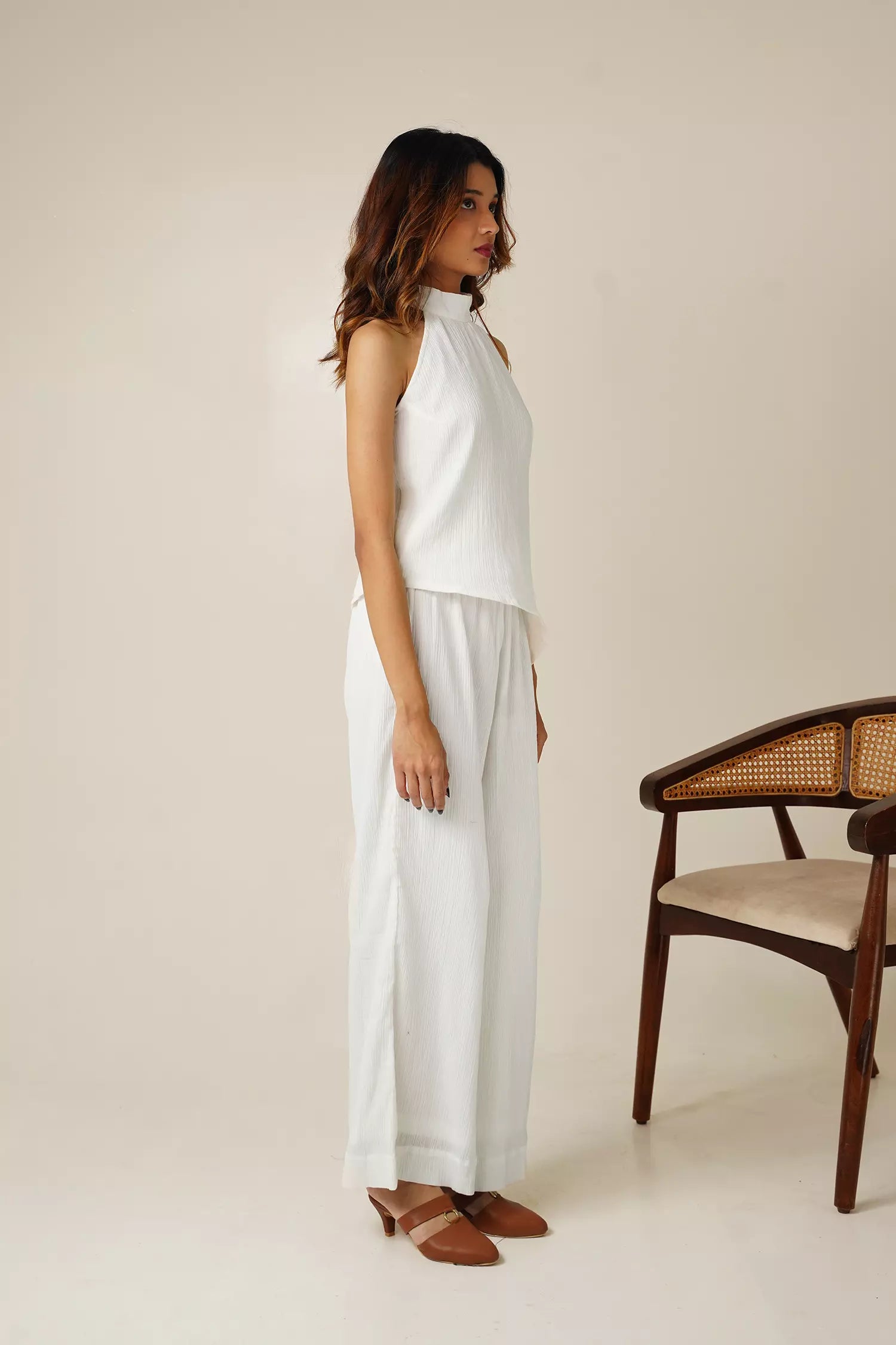side view of Woman wearing a white crinkle cotton halter neck sleeveless top and wide-leg pants standing against a beige background with a wooden chair.