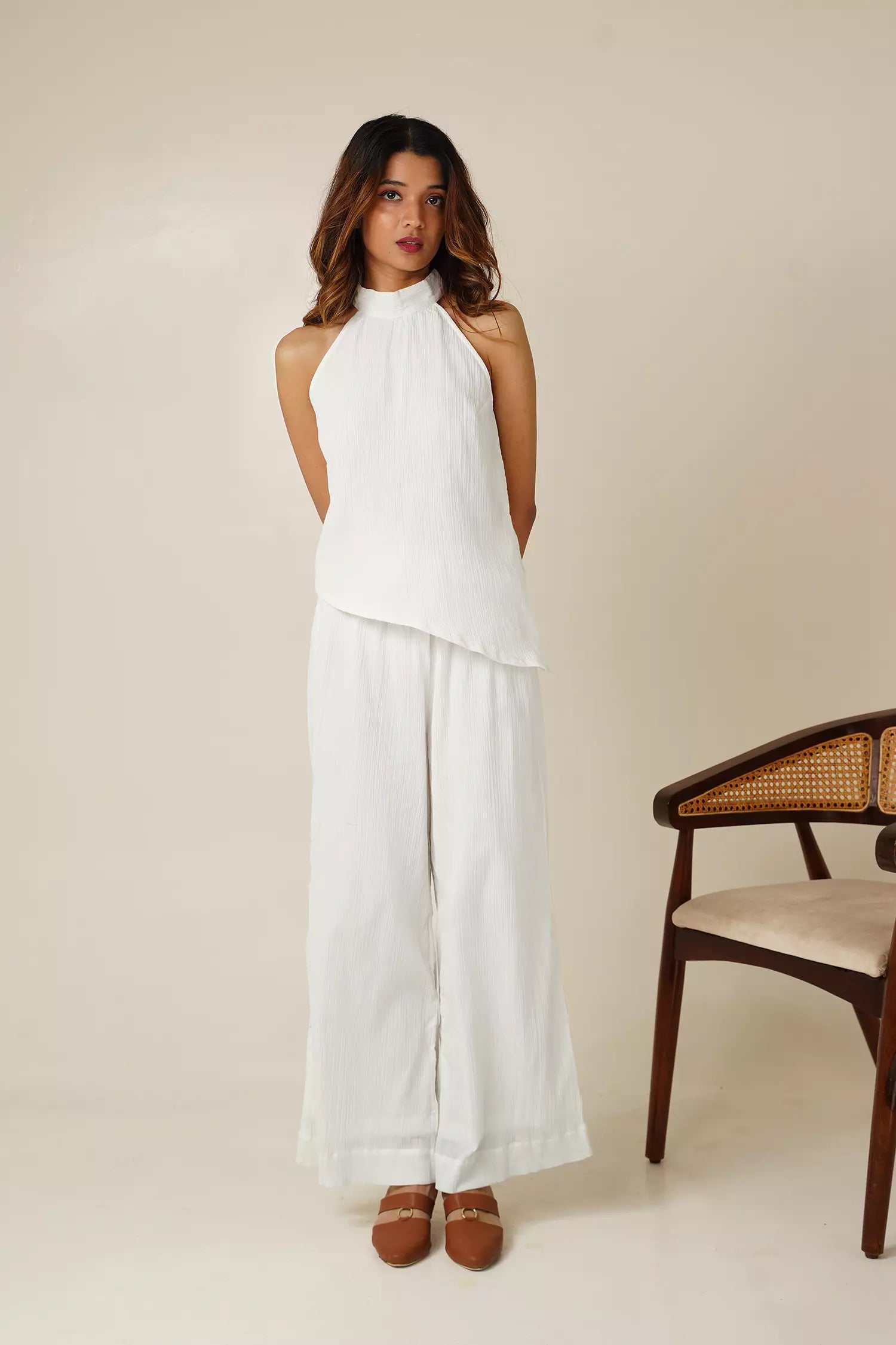 front view of Woman wearing a white crinkle cotton halter neck sleeveless top and wide-leg pants standing against a beige background with a wooden chair.