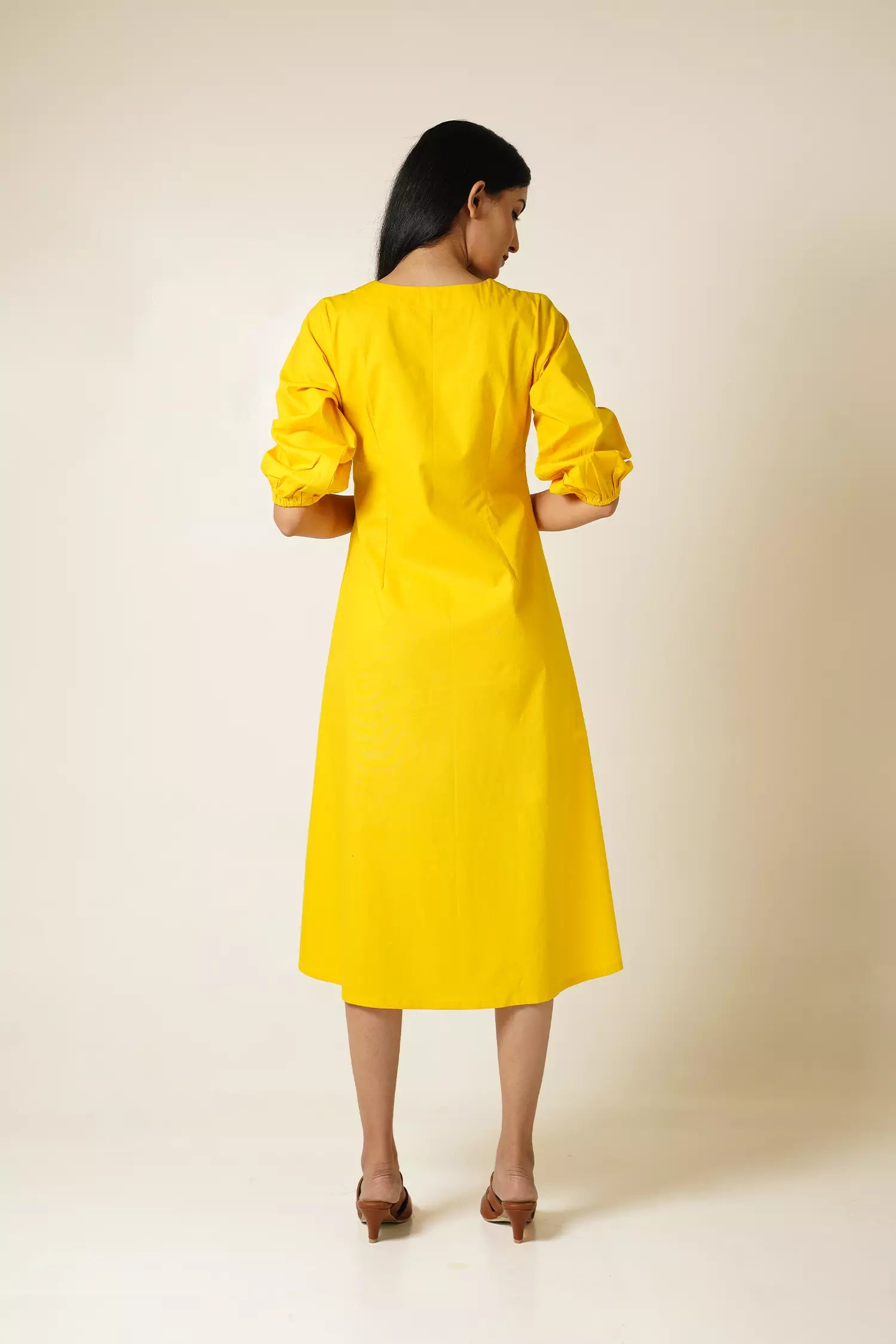 back view of a Woman wearing a cotton midi length yellow dress with v neck and puff sleeves against a plain background