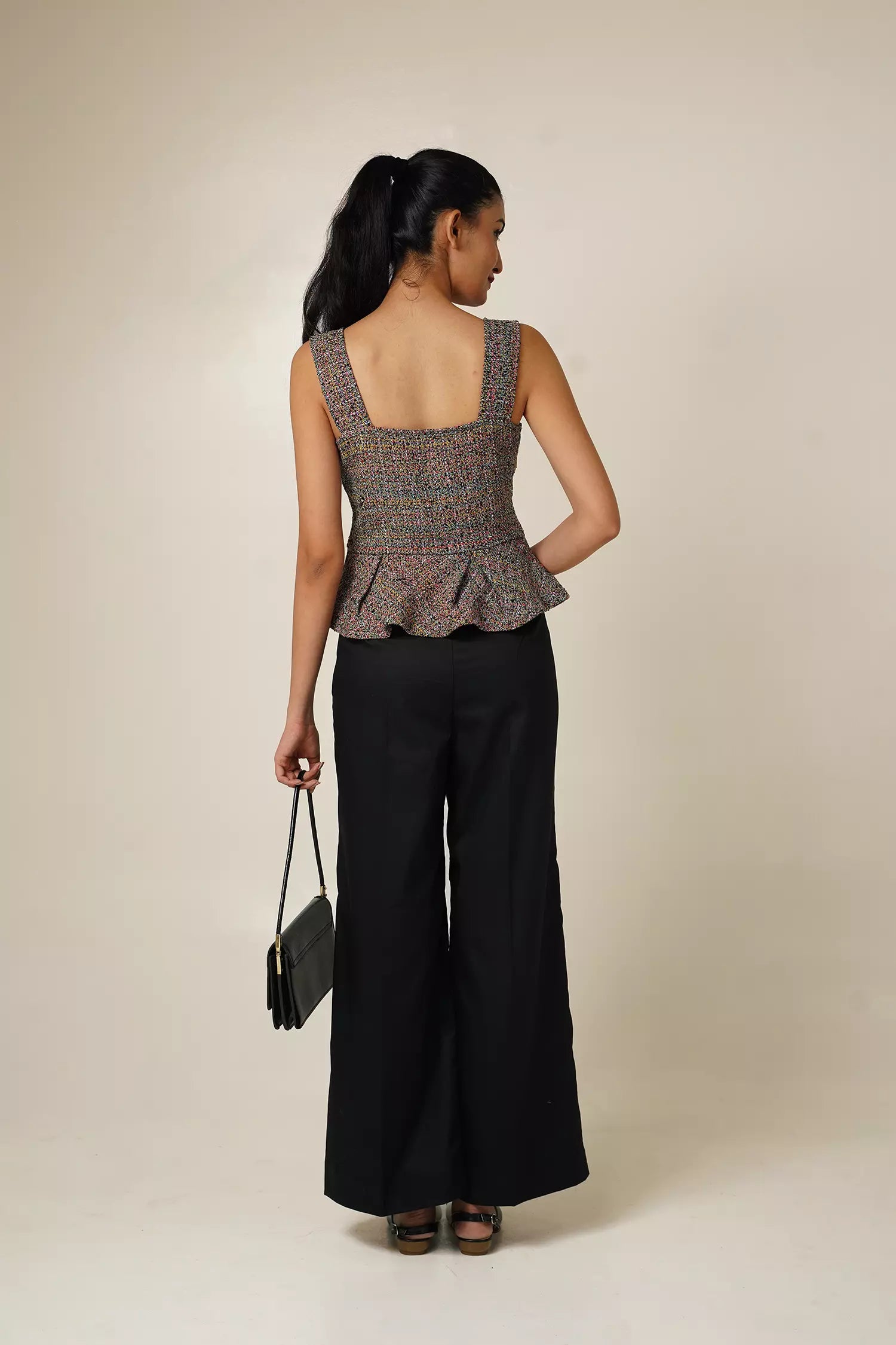 back view of Woman wearing a multi coloured sleeveless tweed top with square neckline and peplum bottom and black pants with a beige background