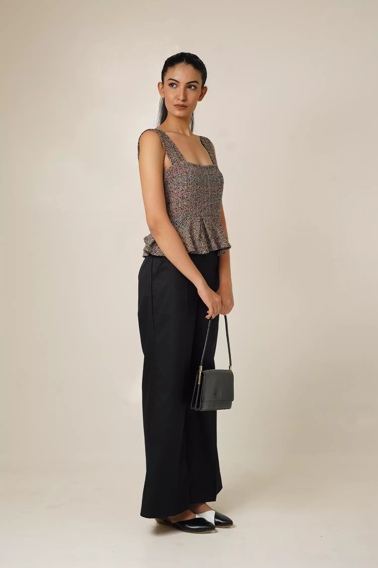 side view of Woman wearing a multi coloured sleeveless tweed top with square neckline and peplum bottom and black pants with a beige background