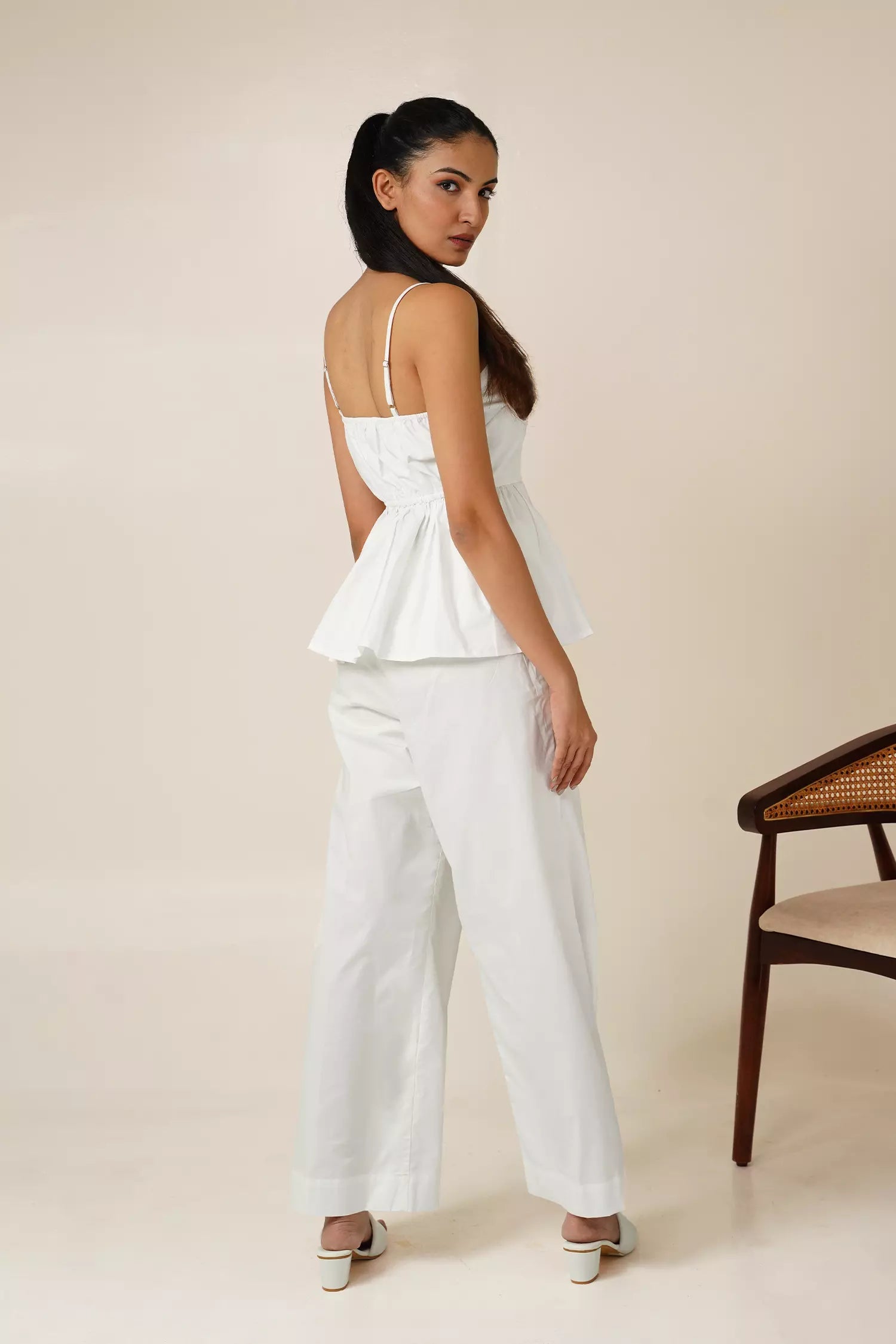 back view of Woman wearing a white odd lama co-ord set of sleeveless peplum top and wide fit pants in pure cotton, holding a brown bag against a beige background