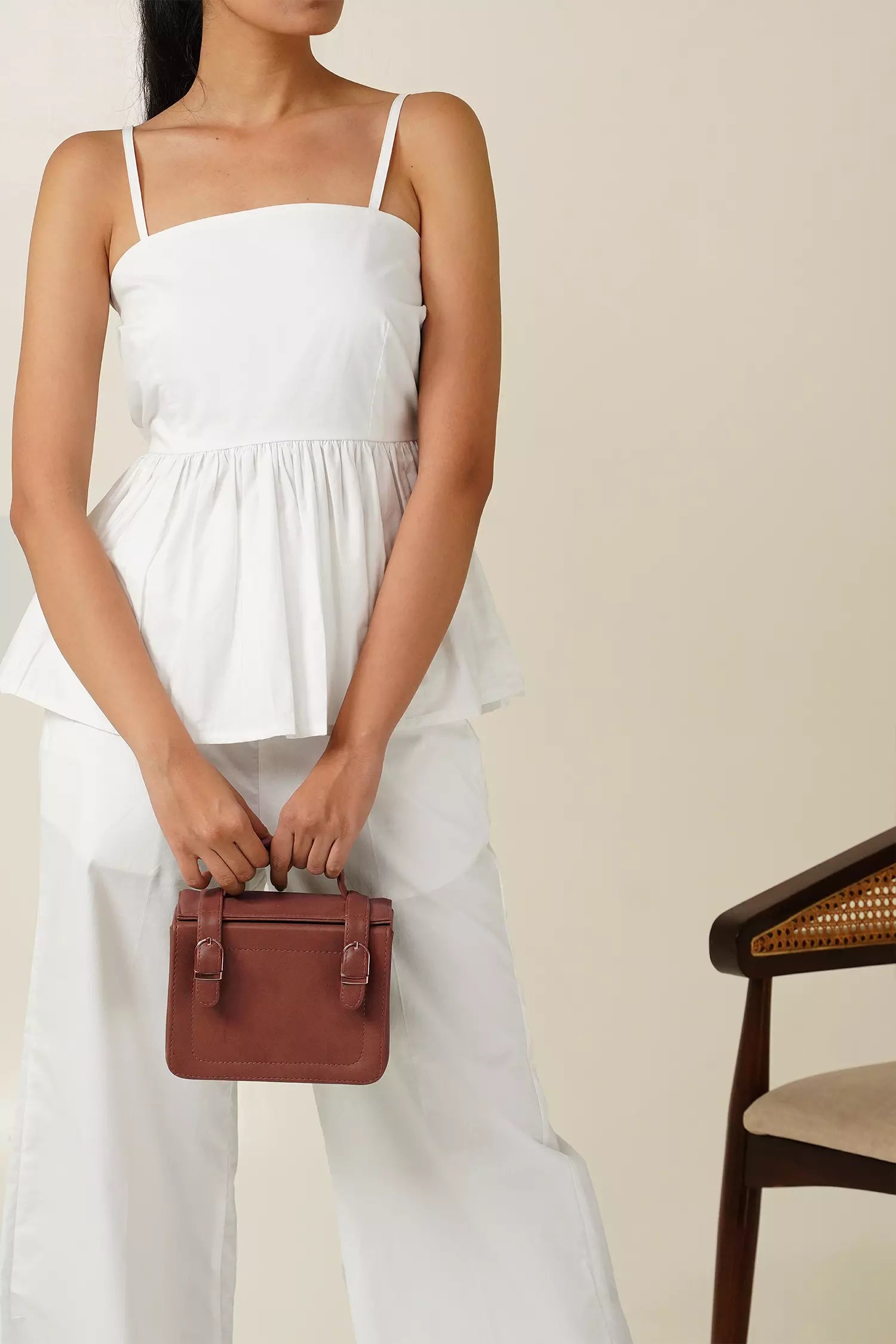 close up front view of Woman wearing a white odd lama co-ord set of sleeveless peplum top and wide fit pants in pure cotton, holding a brown bag against a beige background