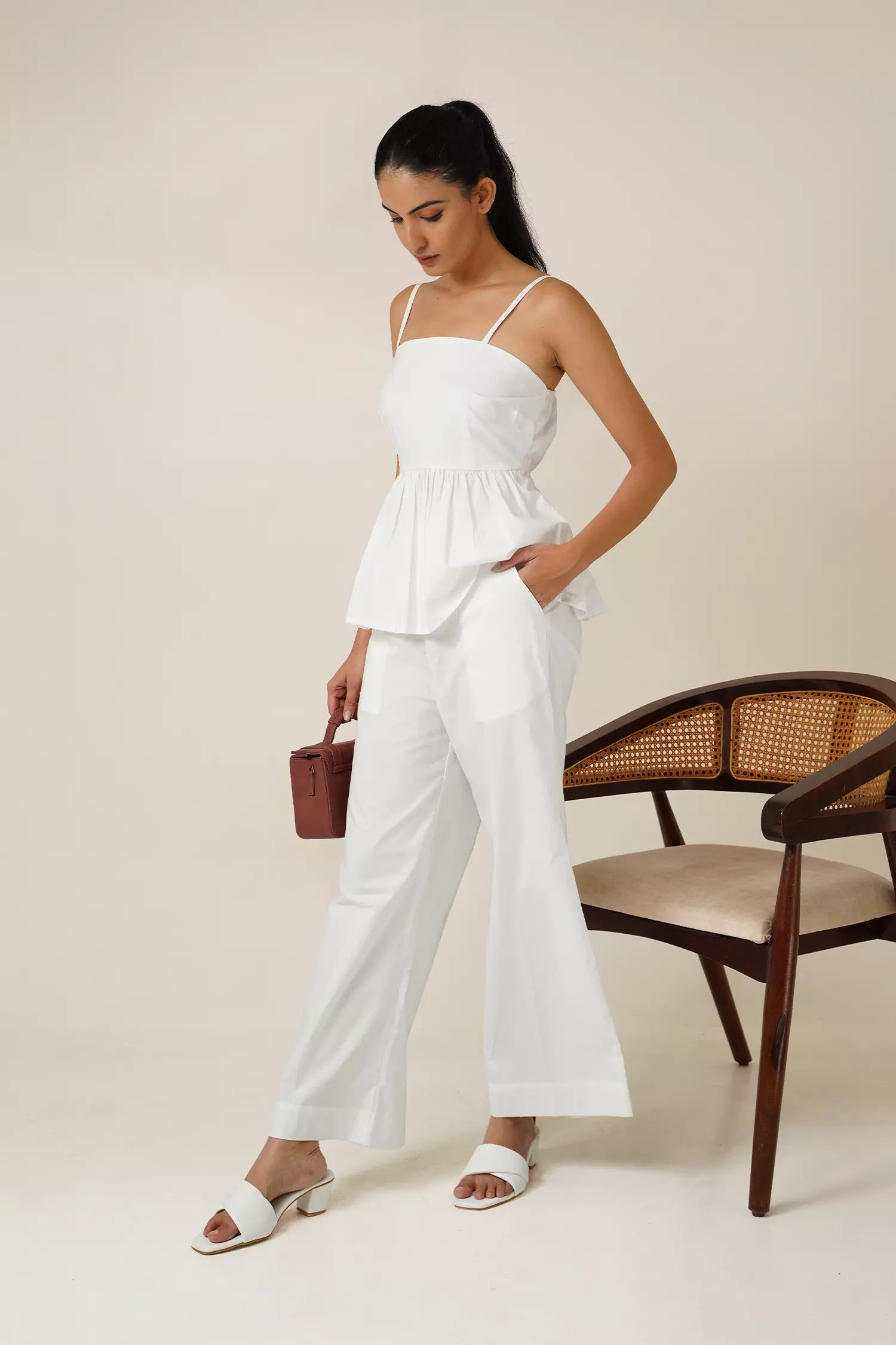 side view of Woman wearing a white odd lama co-ord set of sleeveless peplum top and wide fit pants in pure cotton, holding a brown bag against a beige background