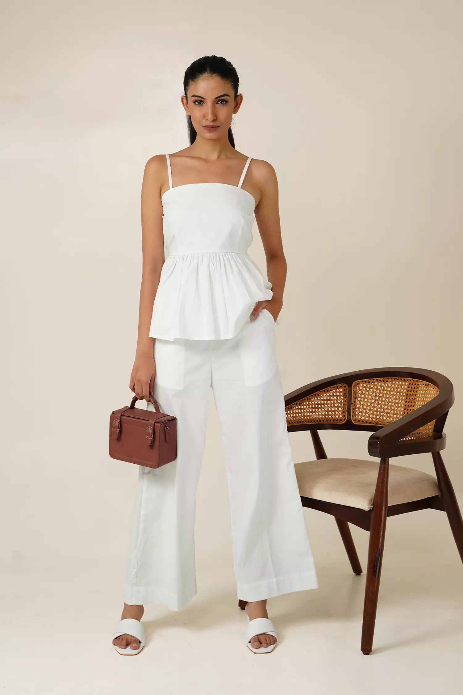 front view of Woman wearing a white odd lama co-ord set of sleeveless peplum top and wide fit pants in pure cotton, holding a brown bag against a beige background