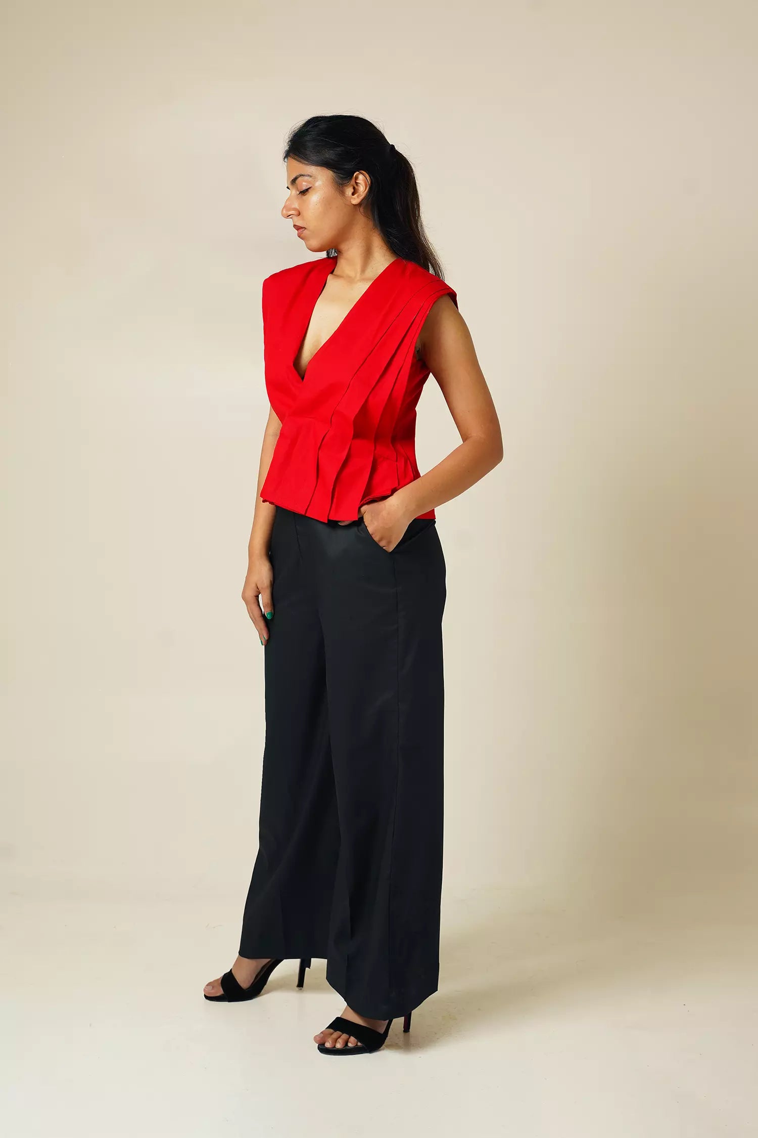 side view of Woman wearing a red pure cotton pleated top with plunging neckline and black pants against a beige background