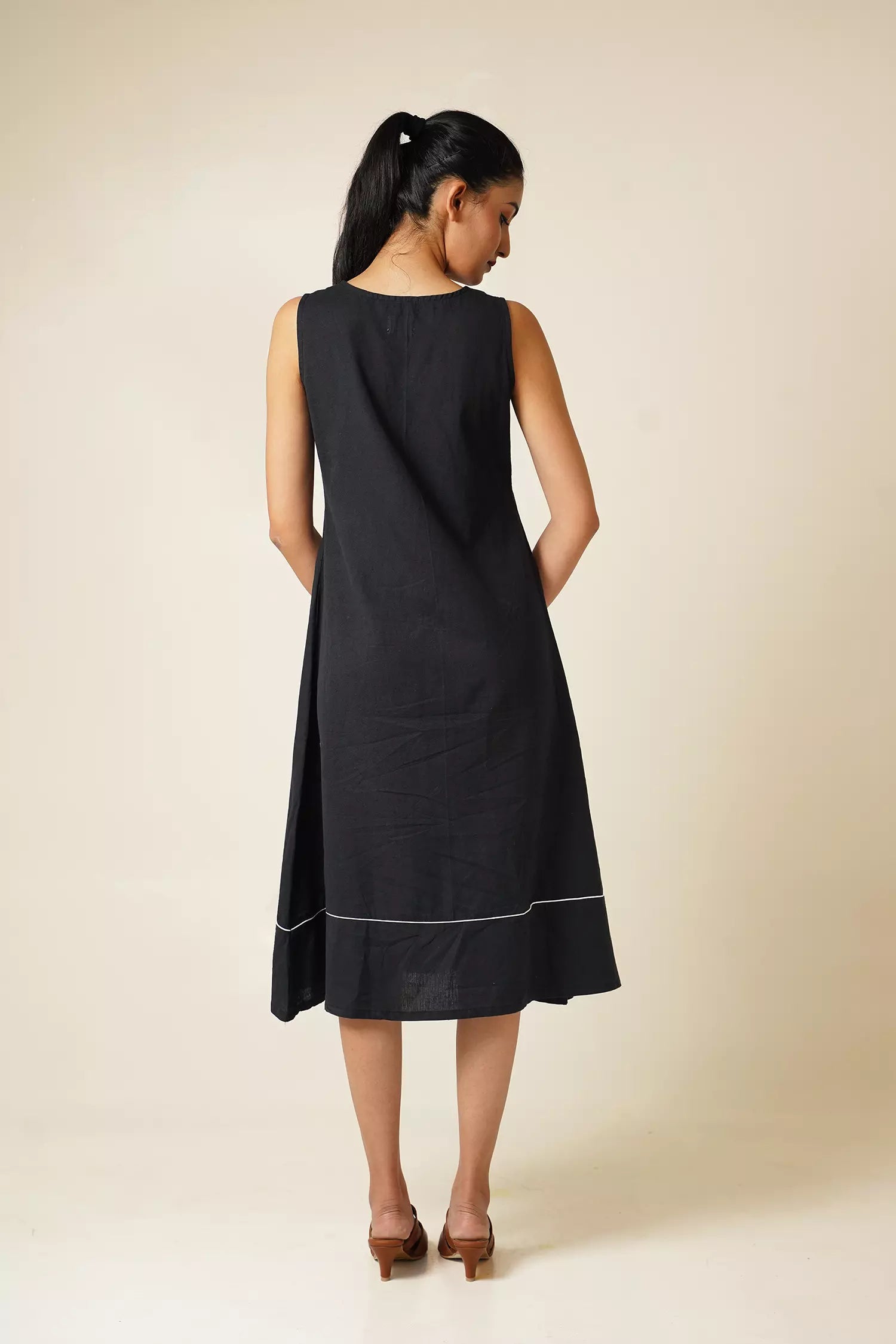 back view of Woman wearing a black knee length sleeveless dress with modified v neckline on a beige background