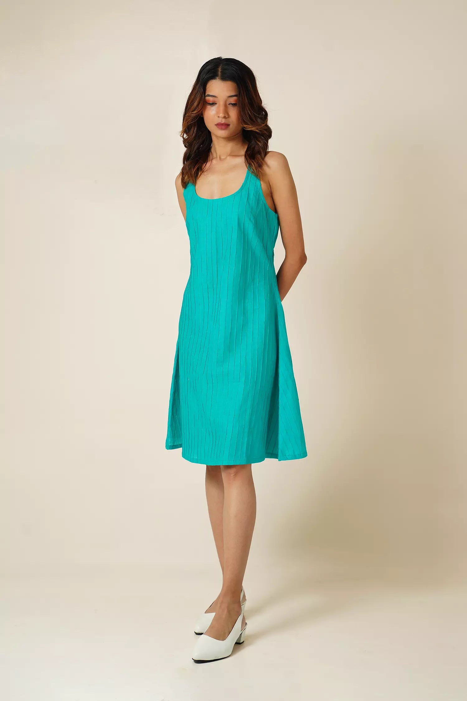 front view of Woman wearing a teal sleeveless knee length  dress with pin tucks on cotton fabric against a beige background