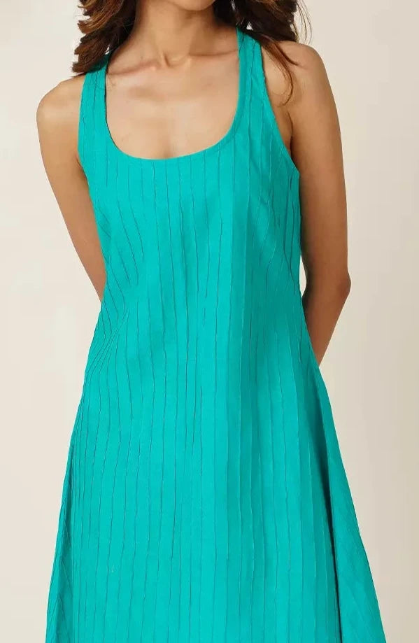 front view of Woman wearing a teal sleeveless knee length  dress with pin tucks on cotton fabric against a beige background