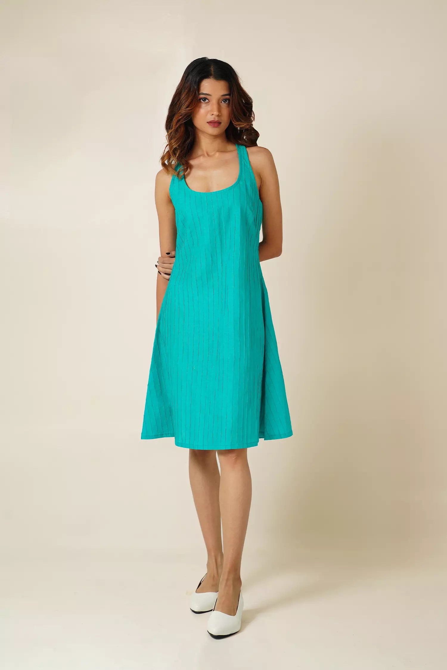 front view of Woman wearing a teal sleeveless knee length  dress with pin tucks on cotton fabric against a beige background