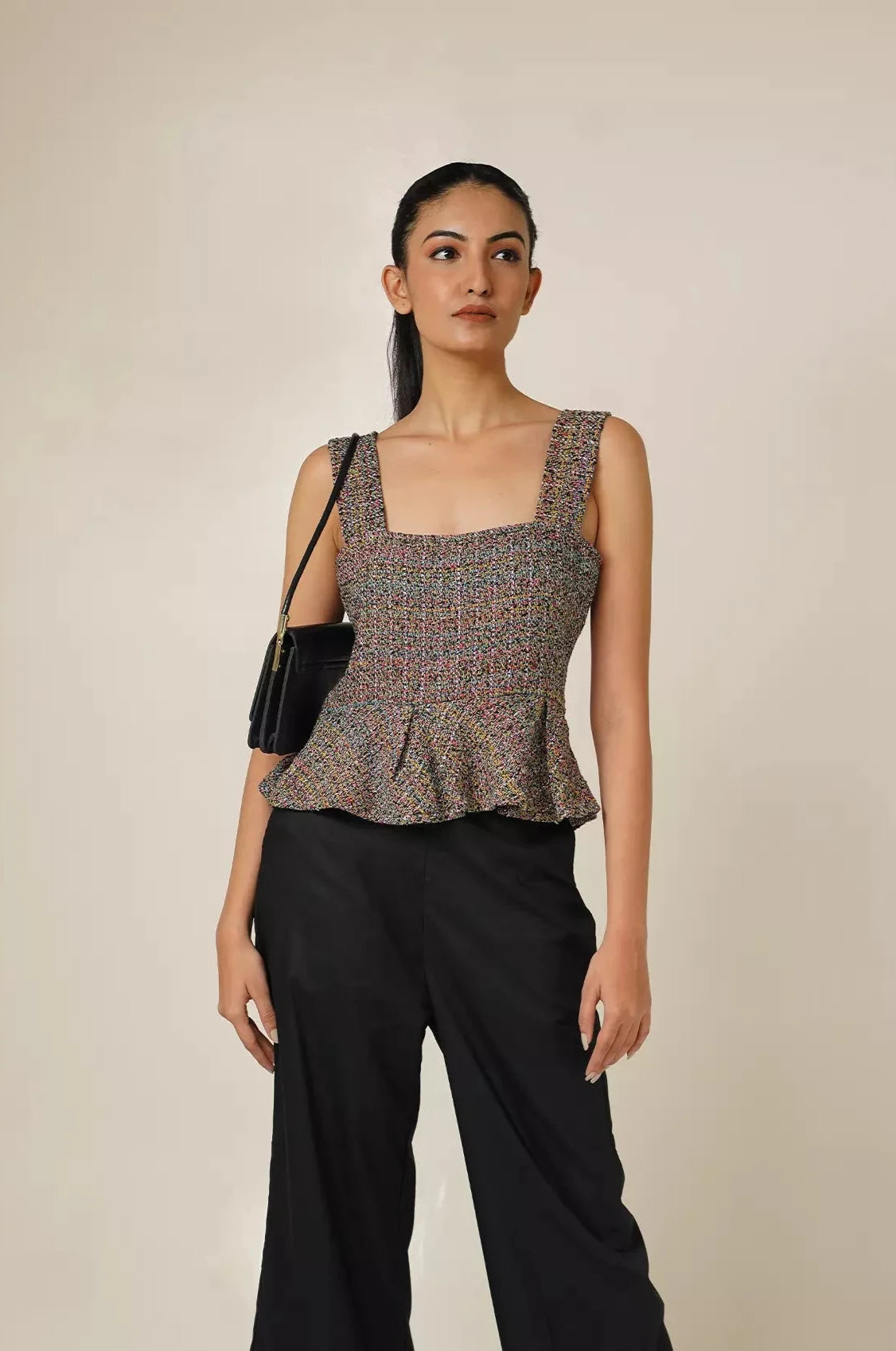 front view of Woman wearing a multi coloured sleeveless tweed top with square neckline and peplum bottom and black pants with a beige background